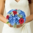 A bridal bouquet that reads like a small, perfectly pitched poem: dense clusters of cerulean and sky-blue hydrangea form a plush, cloud-like cradle whose rounded petals look almost dewy under soft natural light. Tucked into that cool blue haven are several deep red roses, their velvety spirals unfurling with the confident bloom of long-held affection; each rose feels like a heartbeat against the pale lace of a bride's gown. Scattered through the composition, fine sprays of white gypsophila float like whispers of confetti, their tiny stars adding air and scale so the arrangement breathes and moves with every step. Interlaced eucalyptus leaves bring a muted, silvery-green contrast, their matte sheen and subtle, resinous scent grounding the bouquet with a fresh woodland note that calms the senses. The overall palette-blue, scarlet, ivory, and soft green-reads both classic and modern, lit gently so colours glow without glare, texture and fragrance combining into a tactile memoir. As a local florist I picture this carried along Dollis Hill or across the stepped paths by Hendon Park, petals catching sunlight like memories held close. The bouquet is balanced so it sits comfortably in the hands, weighted by blooms but airy at the edges, designed to frame the bride's dress rather than overwhelm it. It is an heirloom-in-waiting that promises ceremony and joy: the cool serenity of hydrangeas, the ardour of roses, the innocence of gypsophila, and the quiet, woodland breath of eucalyptus-an intimate, sensory companion for vows and photographs on a bright Brent Cross day.